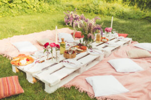 Beautiful summer celebration picnic in the garden with tasty food, cold drinks. Pallet table, Flowers decoration. Pleasure time with friends. Leisure and fun. White and pink colors