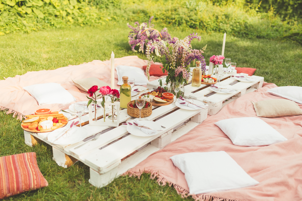 Beautiful summer celebration picnic in the garden with tasty food, cold drinks. Pallet table, Flowers decoration. Pleasure time with friends. Leisure and fun. White and pink colors
