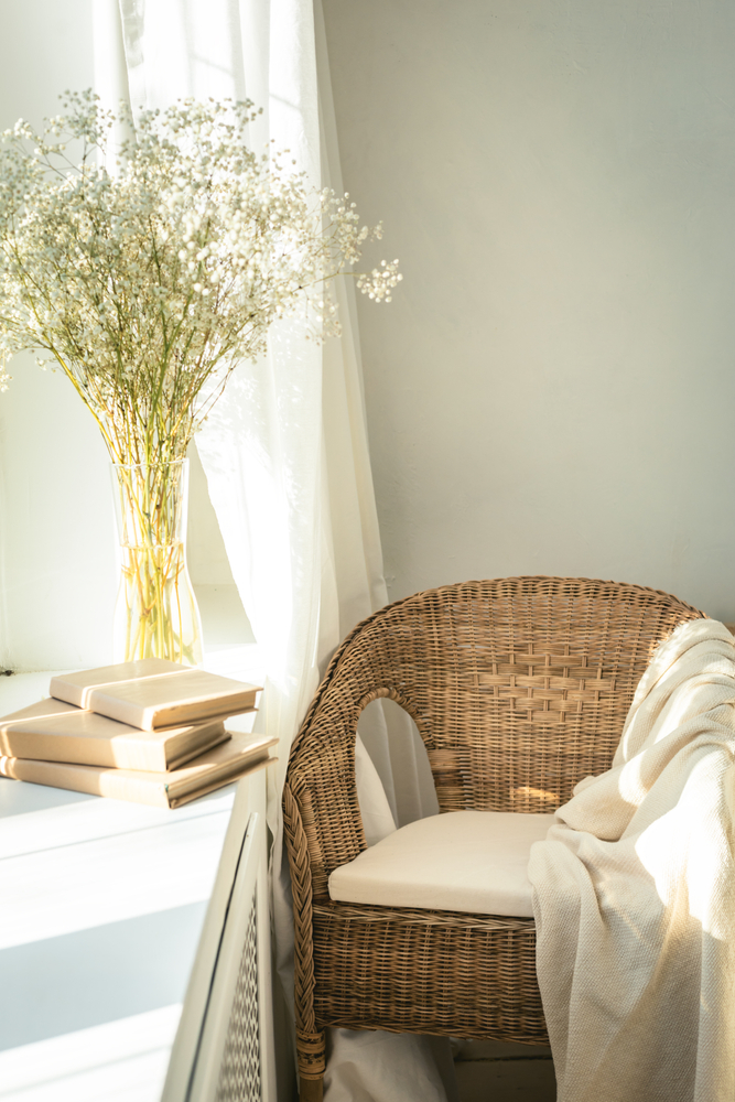 Cozy reading nook by the window with rattan wicker chair, delicate white gypsophila flowers, opened book and window light and shadows.Modern comfortable living room space. Slow living concept
