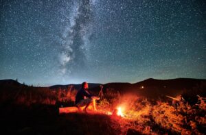 Man taking photos of starry sky with Milky way. Male photographer sitting on log and focusing camera. Man with camera sitting next to camp fire in mountains.