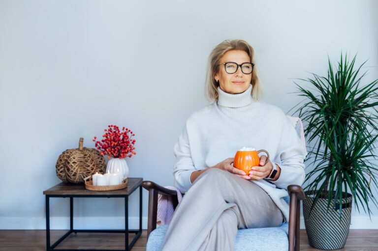 Middle aged woman relaxing with pumpkin shaped cup of hot drink in scandy style hygge interior home with fall mood decor. Lady dreaming, enjoy calm mood without stress, well being alone. Cozy autumn