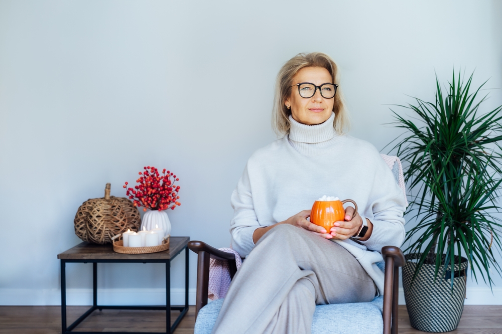 Middle aged woman relaxing with pumpkin shaped cup of hot drink in scandy style hygge interior home with fall mood decor. Lady dreaming, enjoy calm mood without stress, well being alone. Cozy autumn