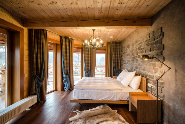 Luxury empty bedroom at home, hotel. Wooden walls animal skin on floor. Lighting equipment hang on ceiling. Bed with clean, comfortable bedding blanket soft pillows. Country landscape through window