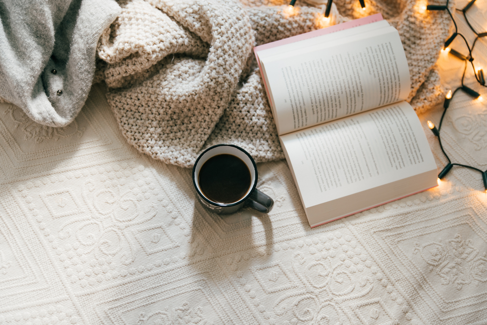 Top view of cozy blanket, open book and hot cup of coffee on bed on a cold winter day. Relaxation and hygge concept. Top view with copy space